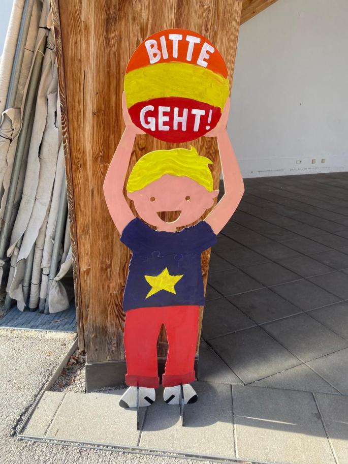 Auf dem Bild sieht man ein Kind aus Holz, welches den Schriftzug "Bitte geht!" trägt.