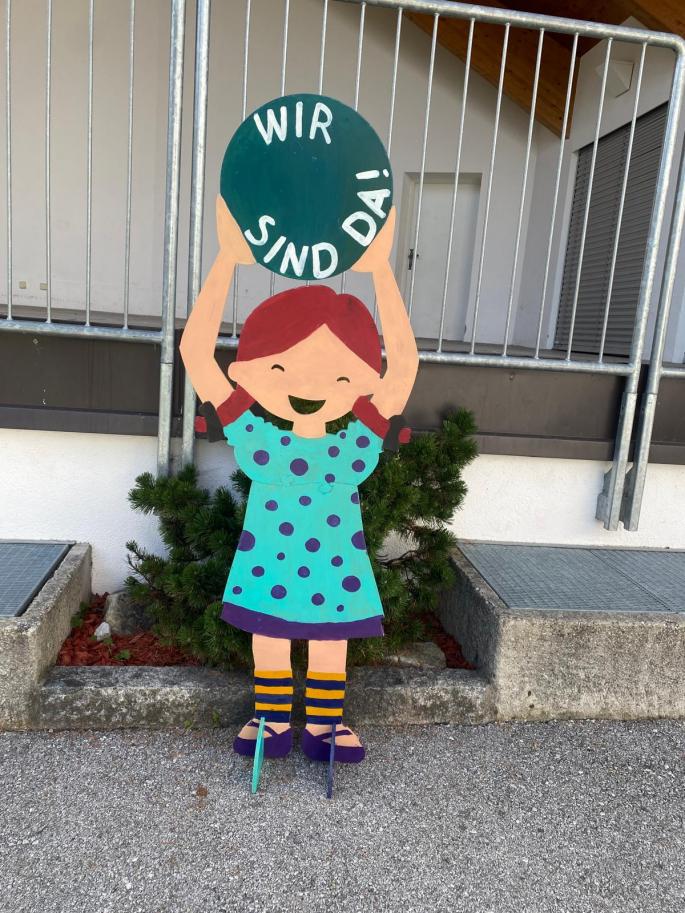 Auf dem Bild sieht man ein Mädchen aus Holz, dass ein Schild mit den Worten "Wir sind da!" trägt