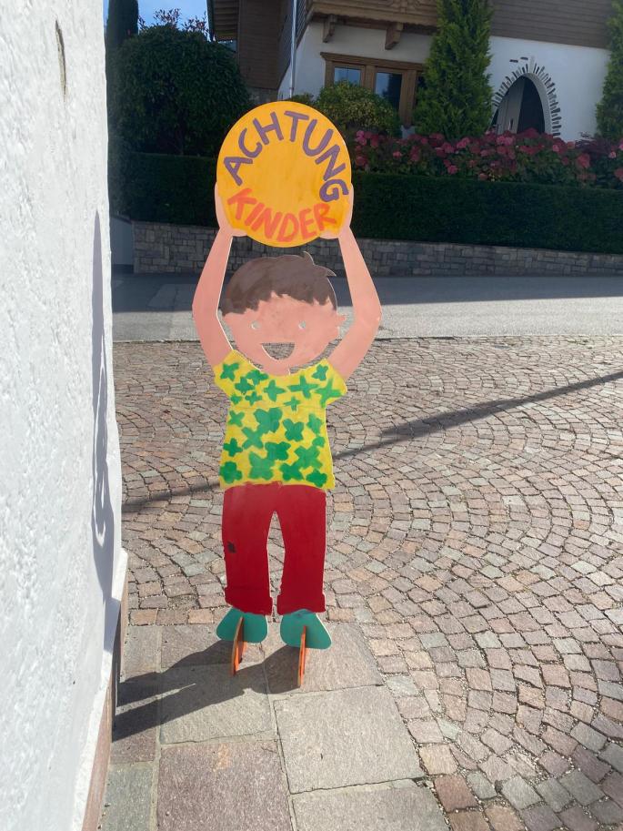 Auf dem Bild sieht man einen Jungen aus Holz, der ein Schild mit den Worten "Achtung Kinder!" trägt.